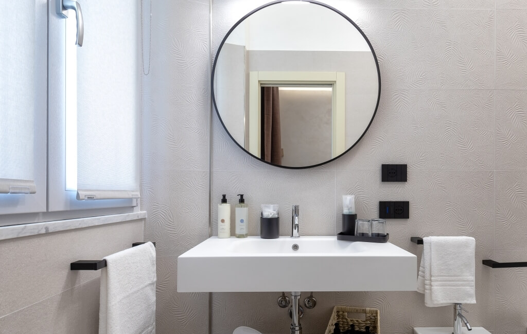 Specchio circolare sopra un lavabo bianco in un bagno luminoso con piastrelle grigie e vari articoli da toeletta.