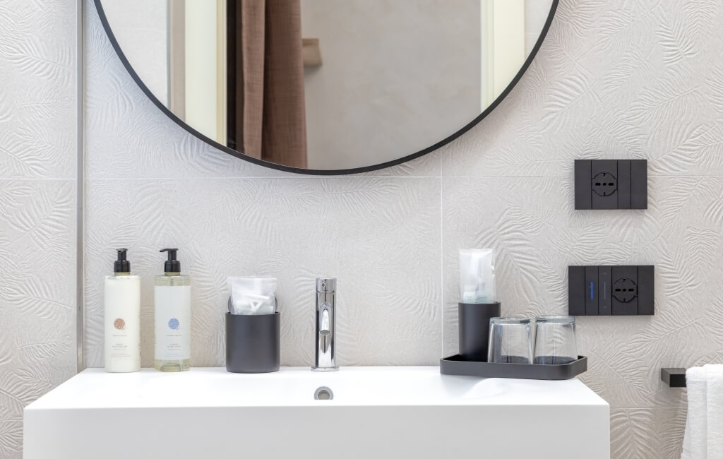 Area lavabo moderna con lavabo bianco, specchio rotondo, e due flaconi di sapone su un piano bianco.