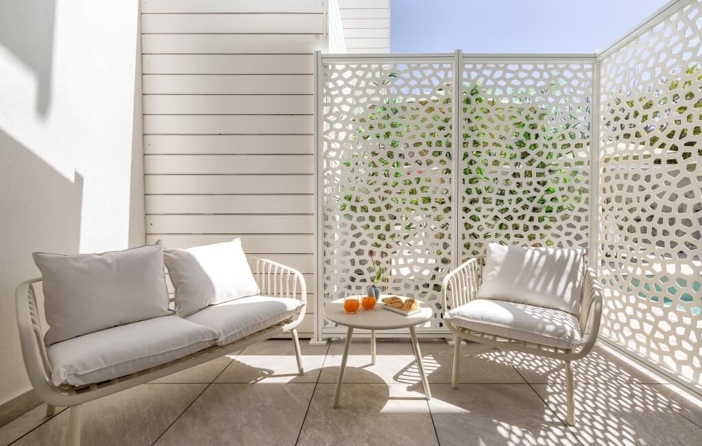 Area esterna serena con mobili bianchi, schermo a graticcio e bevande arancioni su un tavolo, immersa nella luce naturale.