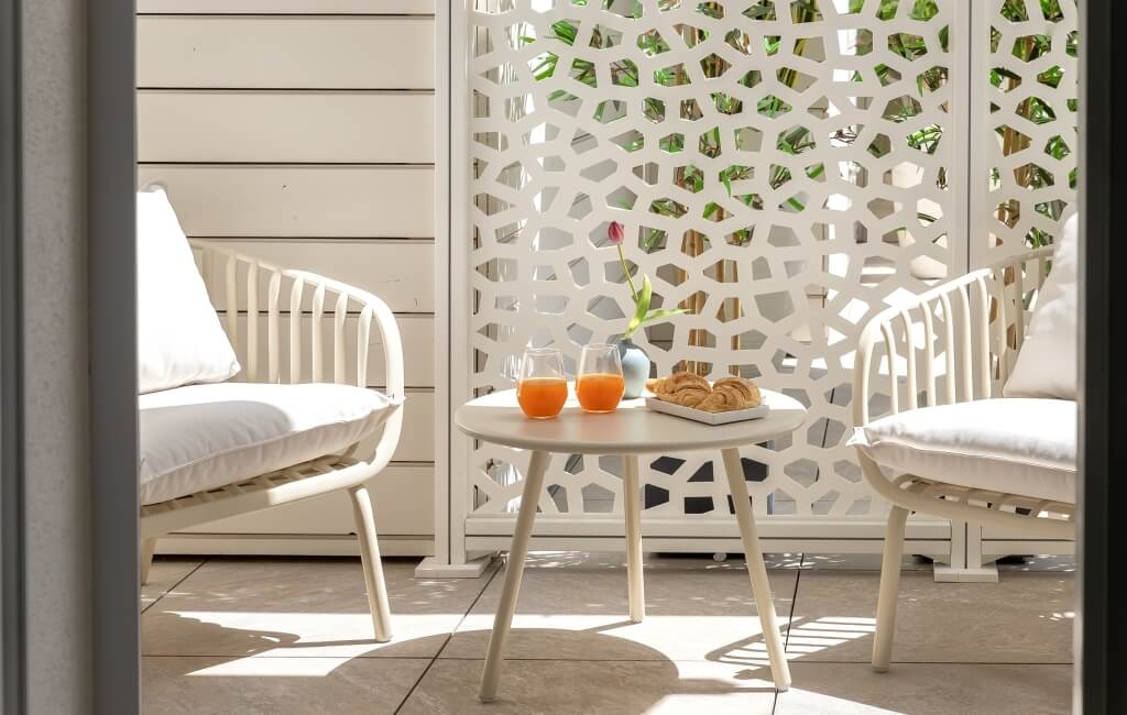 Tavolo bianco rotondo con croissant e succhi d'arancia su un patio soleggiato, circondato da due sedie bianche.