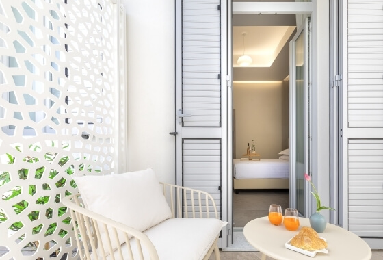 Area esterna con sedia bianca, tavolino e vista su una camera da letto elegante con letto bianco e illuminazione soffusa.