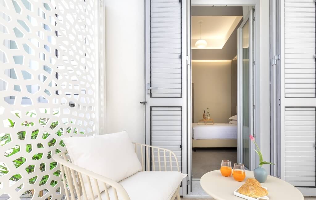 Area esterna rilassante con sedia bianca, tavolo rotondo con colazione e vista su una camera da letto attraverso una porta aperta.