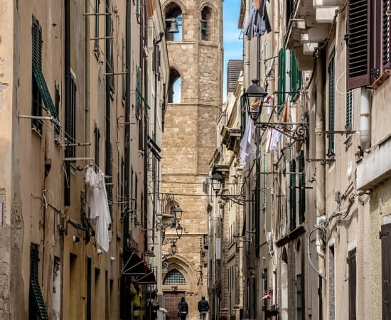 Vicolo stretto tra edifici in pietra chiara, con torre di chiesa in lontananza e oggetti appesi alle pareti.