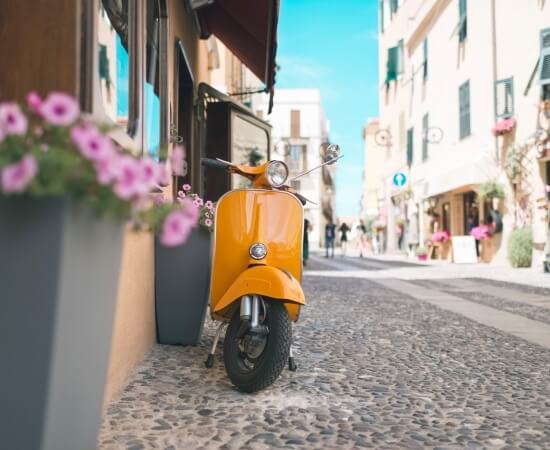 Scooter giallo parcheggiato su una strada acciottolata in una città europea, con fiori rosa e cielo blu sullo sfondo.