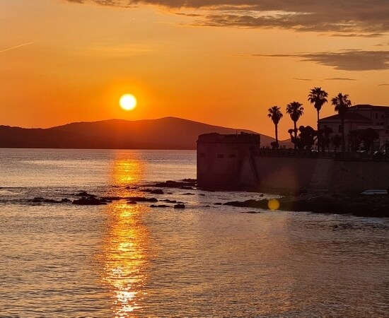 Tramonto sereno su un corpo d'acqua, con palme e montagne scure sullo sfondo, riflessi arancioni nell'acqua calma.