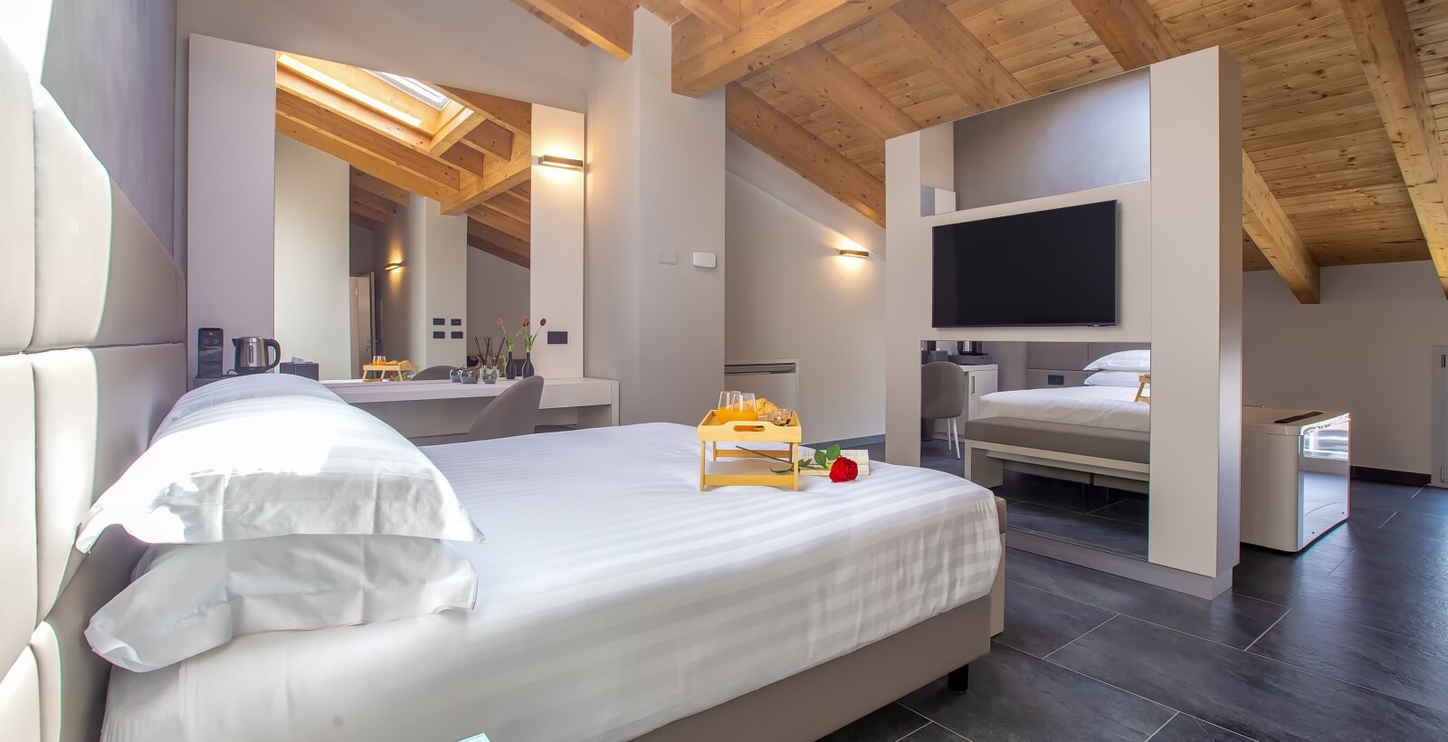 Camera d'hotel con letto bianco, vassoio giallo con succo d'arancia e croissant, illuminata da luce naturale e artificiale.