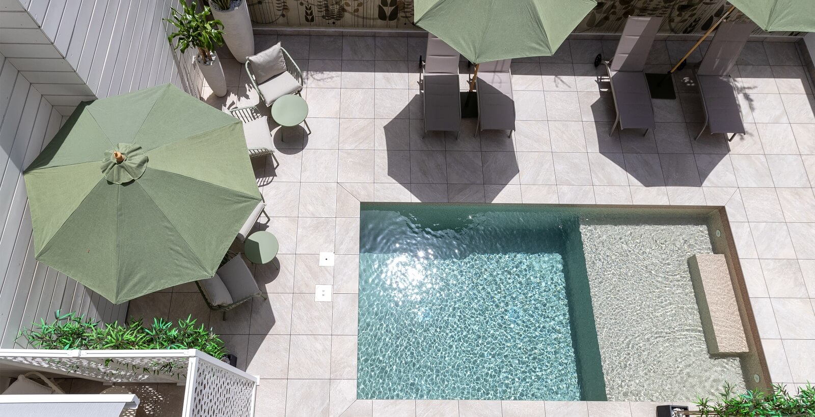 Vista aerea di un patio moderno con piscina, ombrelloni verdi e sedie grigie su piastrelle chiare.
