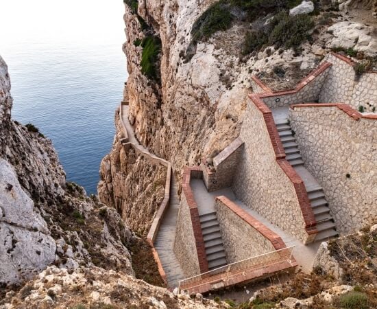 Scala in pietra con bordi in mattoni rossi, incastonata in una scogliera, con vista su un mare blu calmo.