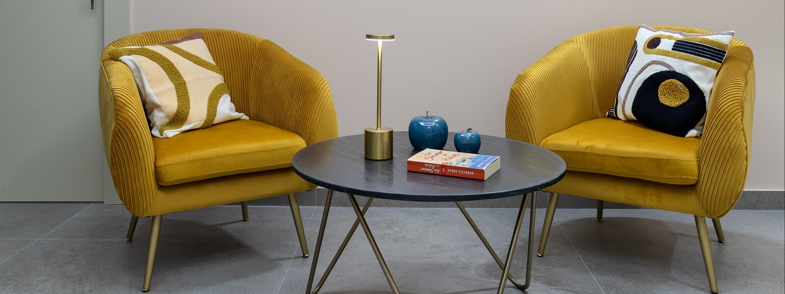 Due poltrone giallo senape su un pavimento grigio, un tavolino grigio scuro con lampada dorata e decorazioni blu.