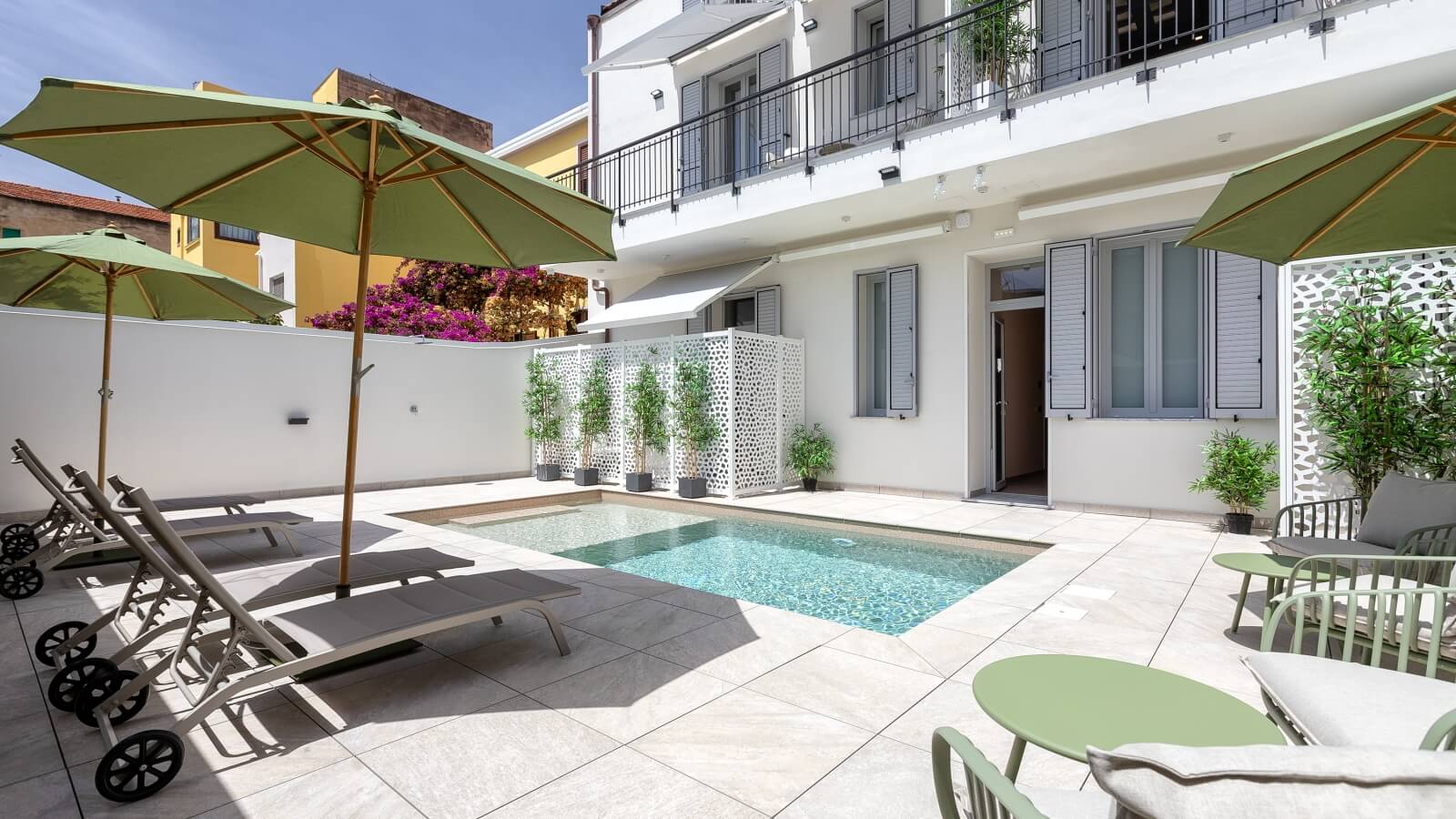 Area patio con piscina, lettini grigi, ombrelloni verdi e piante artificiali, circondata da un edificio bianco e giallo.