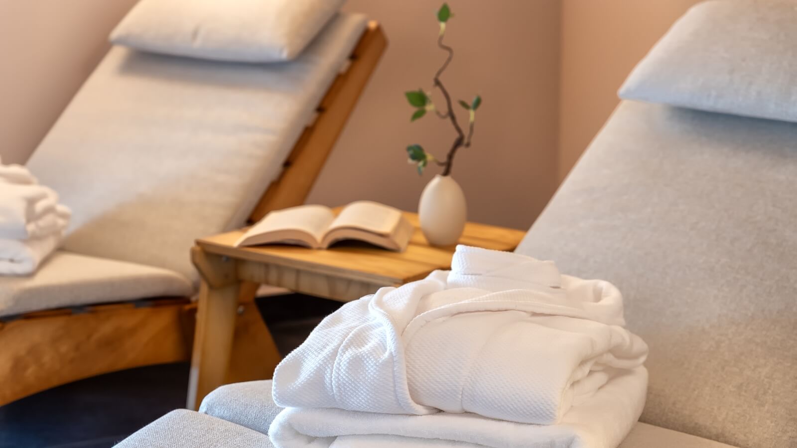 Due lounge chair in un'accogliente spa, con asciugamani bianchi e un tavolino che tiene un libro e una pianta.