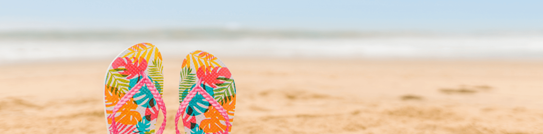 Un paio di infradito colorate con design tropicale in primo piano, con spiaggia e oceano sullo sfondo.