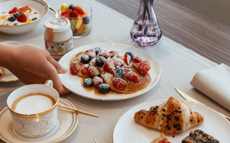 Colazione con pancake, croissant, cereali, frutta e caffè.