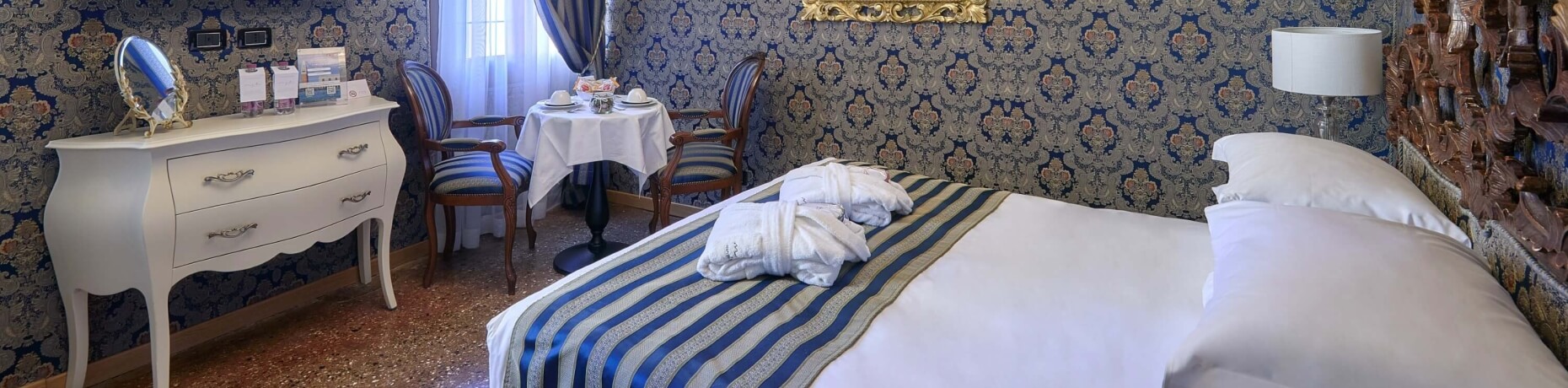 Camera elegante con carta da parati blu e oro, letto con lenzuola bianche, tavolo con colazione e sedie a righe.