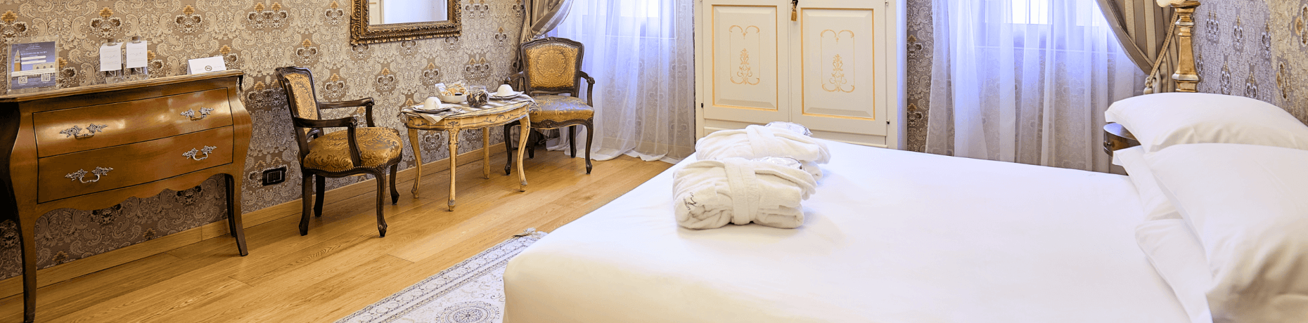 Camera d'hotel con pavimento in legno, mobili scuri, tappeto orientale e luce soffusa che filtra da tende bianche.