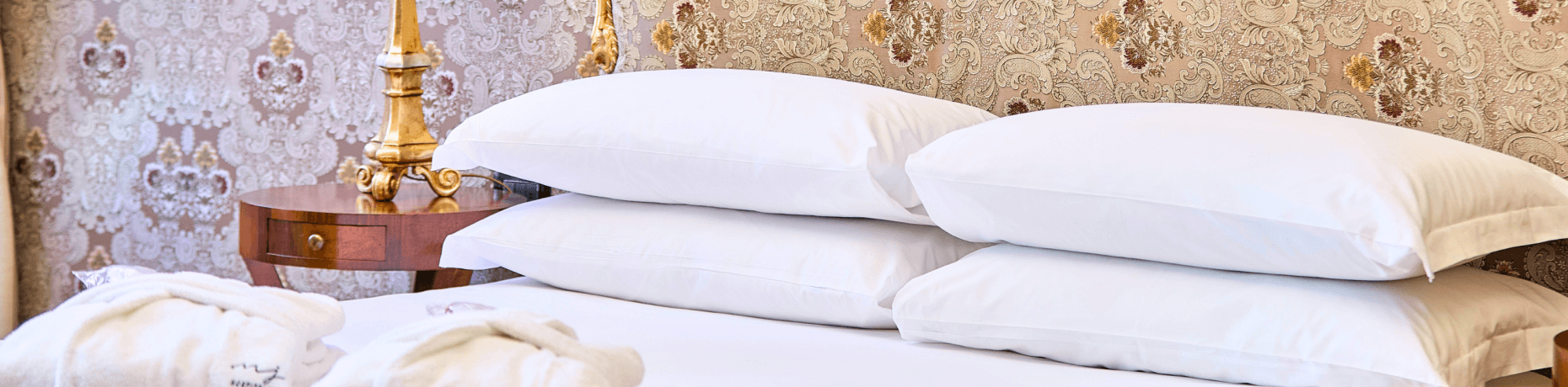 Ein elegantes Bett mit weißen Kissen und Decke, einem Holz-Nachttisch und einer goldenen Lampe vor floralem Tapetenhintergrund.