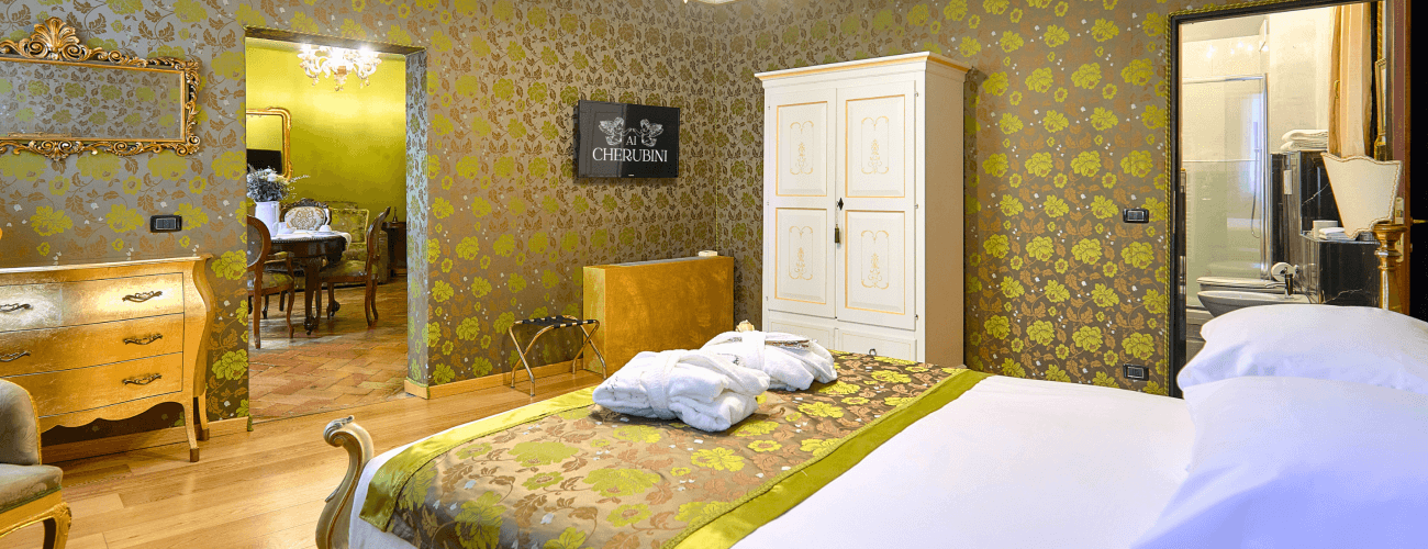 Großes Schlafzimmer mit Blumen-Tapete, Bett mit weißen Decken, Schränken und Zugang zu einem Essbereich mit "ALBERGO CHERUBINI"-Schild.
