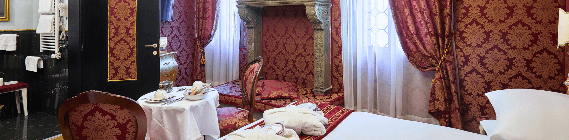 Stanza decorata con tavolo rotondo, tè, poltrona damascata rossa e oro, letto con biancheria bianca e cuscini decorati.
