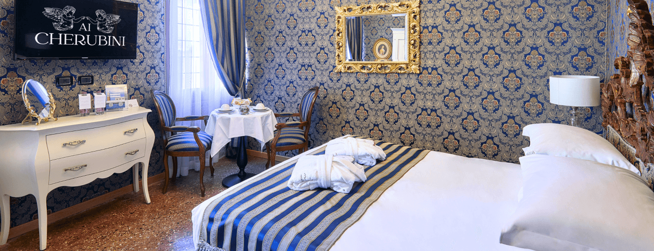 Elegantes Hotelzimmer mit blau-goldenem Dekor, Bett, Kommode, Tisch und Fenster mit weißen Vorhängen.