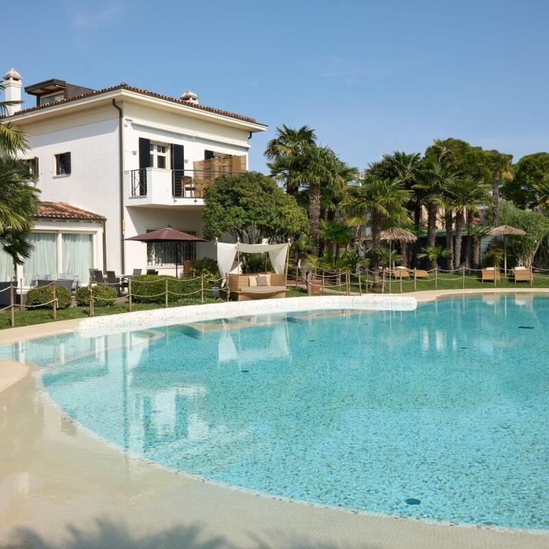 Scène extérieure sereine avec une maison luxueuse, piscine bleue, chaises longues et jardin tropical sous un ciel clair.