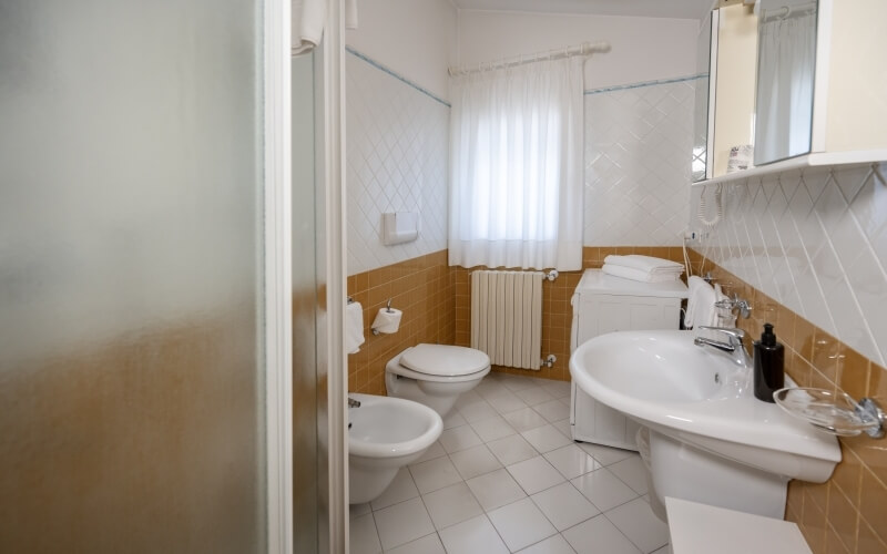 Badezimmer mit weißen und gelben Fliesenwänden, Waschbecken, Toilette, Bidet, Schrank und Fenster mit weißen Vorhängen.