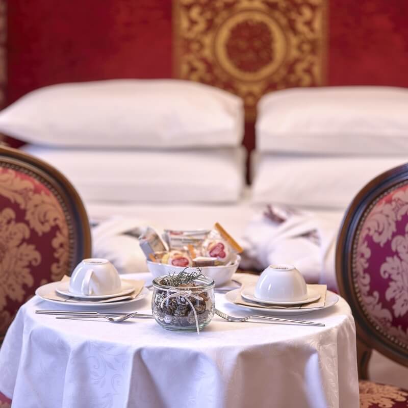 Elegant hotel room table setting for two, featuring white linens, teacups, and decorative items, with a stylish bed backdrop.