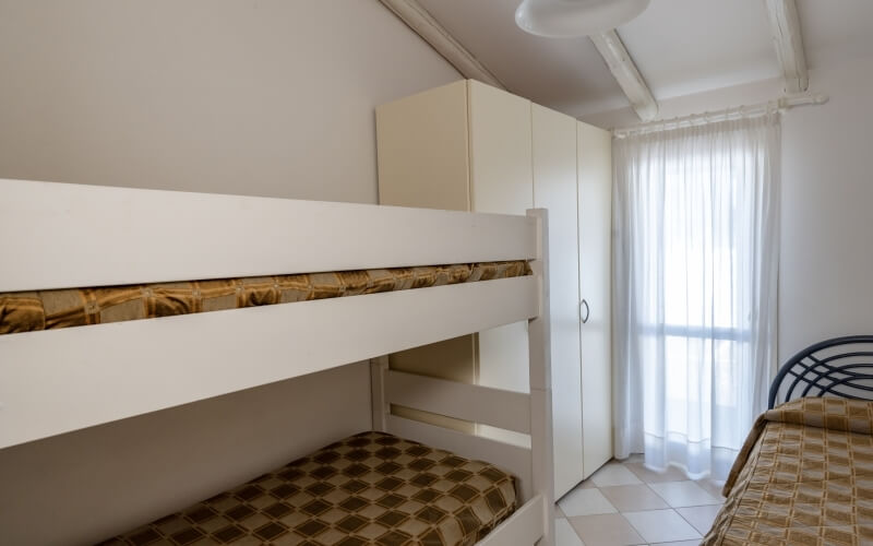 Ein Zimmer mit einem weißen Etagenbett, braun-beigen Bettwäsche, einem weißen Schrank und einem Fenster mit weißem Vorhang.
