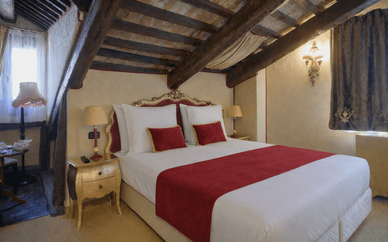 Camera d'hotel con letto bianco, dettagli rossi e travi a vista.