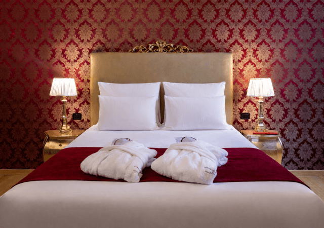 Camera d'hotel con letto bianco, testiera dorata e pareti rosse.