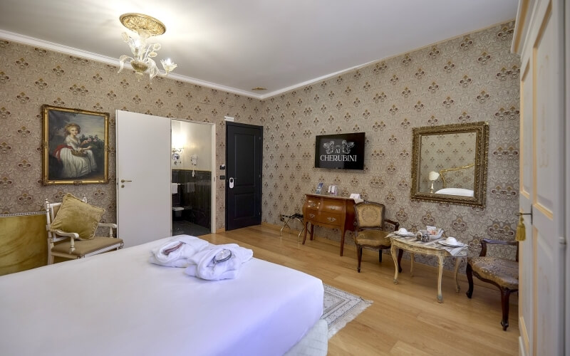 Chambre vintage avec lit, miroir doré, TV, fleurs, et salle de bain, décorée avec papier peint floral et lustre.