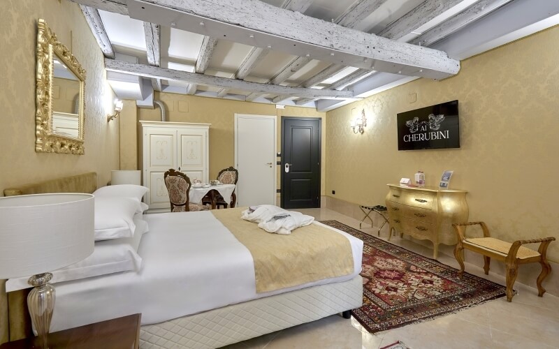 Chambre d'hôtel luxueuse avec lit central, commode en bois clair, tapis rouge et or, et décor classique élégant.