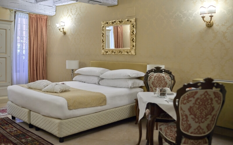 Chambre d'hôtel élégante avec lit, chaises dorées, miroir, fenêtres avec rideaux roses et tapis beige.