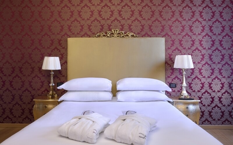 Chambre d'hôtel luxueuse avec un grand lit, têtes de lit dorées, peignoirs blancs et murs en papier peint bordeaux floral.