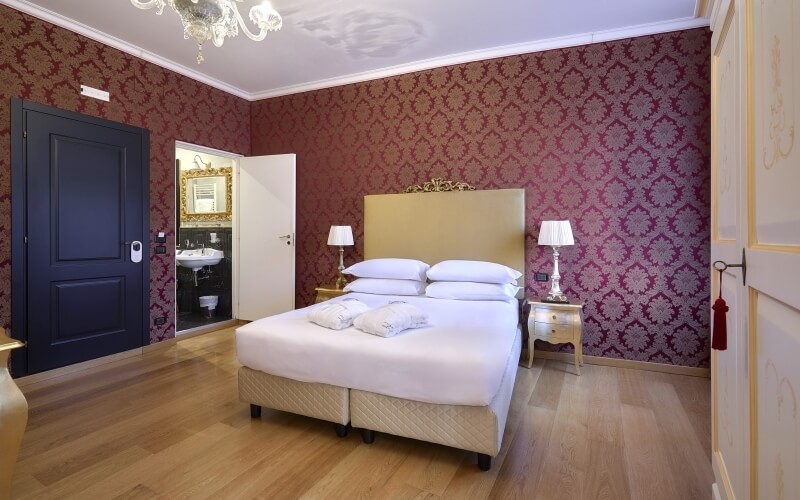 Chambre élégante avec lit central, décor rouge et or, deux tables de nuit, salle de bain et lustre au plafond.