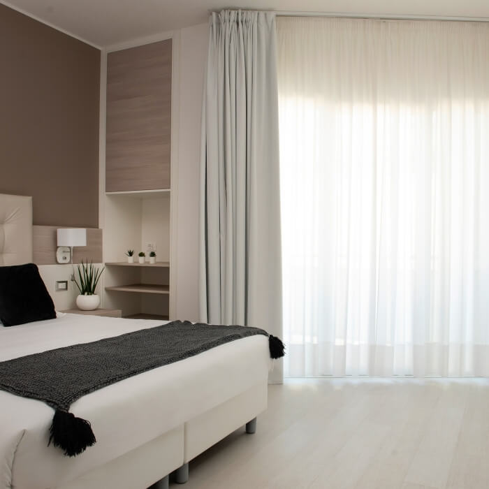 Camera da letto serena con letto bianco, tende leggere, comodino e pareti marroni e bianche. Atmosfera calma e tranquilla.