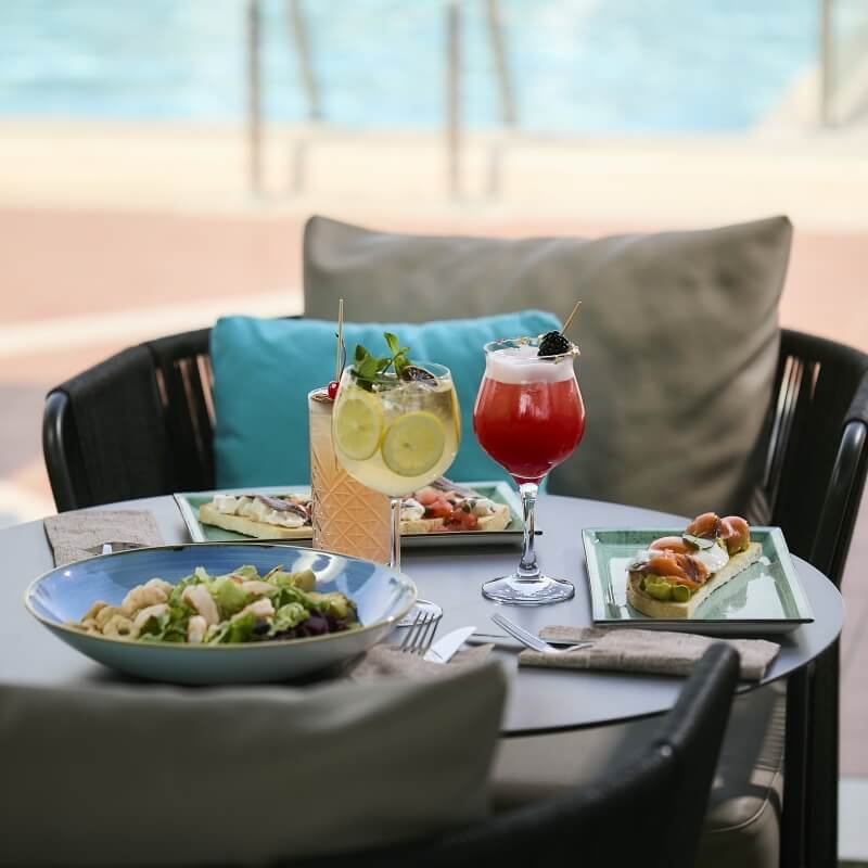 Table ronde avec nourriture et cocktails, piscine en arrière-plan, ciel bleu, ambiance calme et sophistiquée.