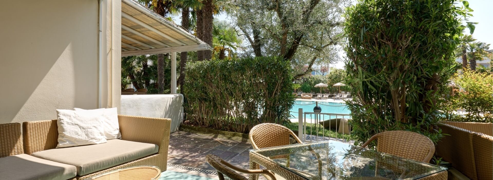 Un patio sereno con divano in rattan, tavolo e sedie, circondato da verde e una piscina, crea un'atmosfera tropicale.