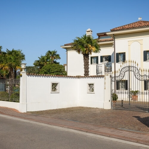 Grande maison blanche avec toit en tuiles, portail en fer forgé, allée en briques, murs blancs et palmiers sous un ciel bleu.