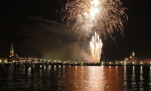 Fuochi d'artificio brillano sopra l'acqua, riflessi luminosi visibili.