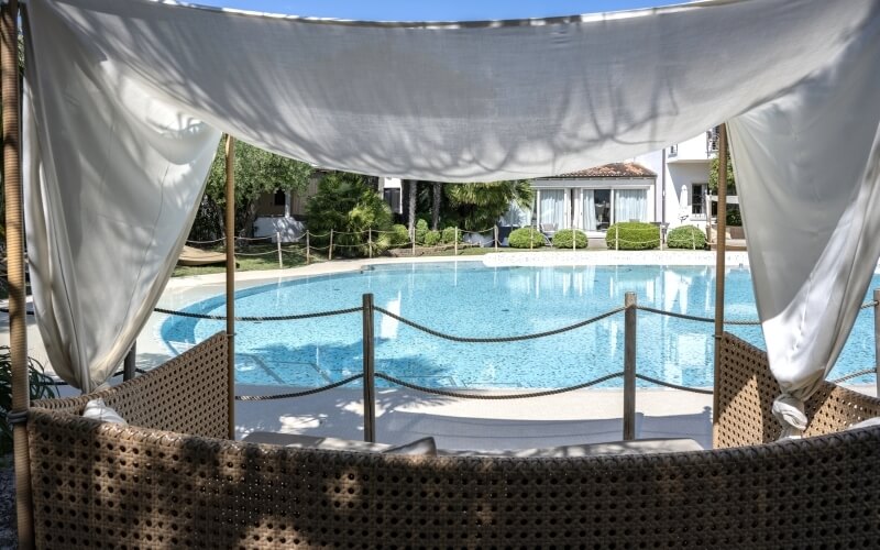 Serene poolside with a rectangular pool, lush greenery, a white house, and shaded wicker chairs under a bamboo canopy.
