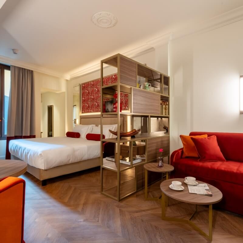 Camera d'hotel con letto bianco, divano rosso e area salotto.