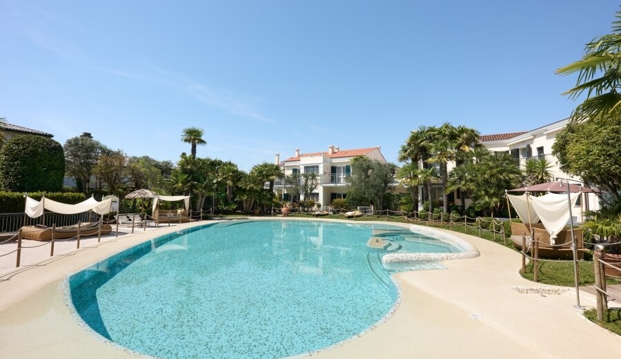 Una piscina con tavoli e chaise lounge, circondata da palme e un complesso residenziale sullo sfondo.