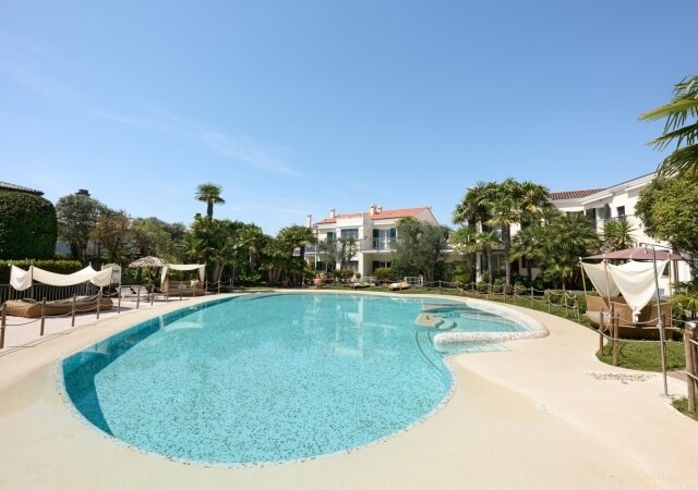 Scena serena di piscina con sedie a sdraio e palme, edifici bianchi sullo sfondo in una giornata di sole.