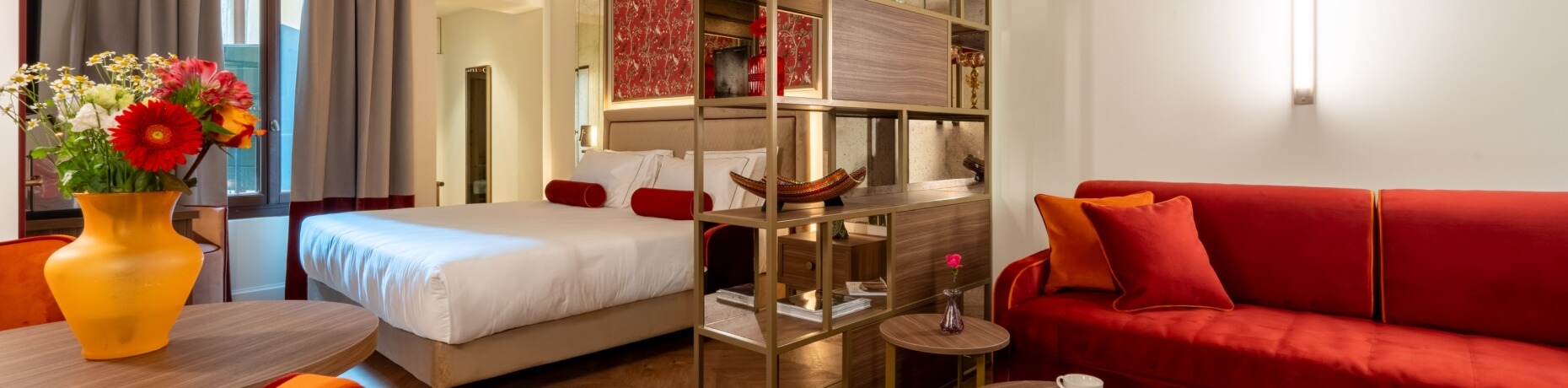 Camera d'hotel con letto, divano rosso e tavolino con fiori.