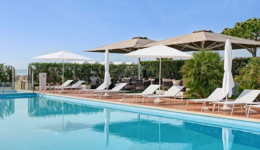 Una scena di un lungo piscina blu con lettini bianchi, ombrelloni e siepi verdi sullo sfondo sotto un cielo azzurro.