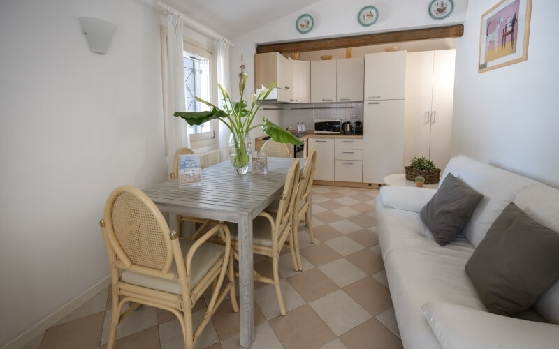 Esszimmer mit grauem Tisch, Rattanstühlen, Pflanzen, Küche im Hintergrund, weiße Vorhänge und dekorative Teller.
