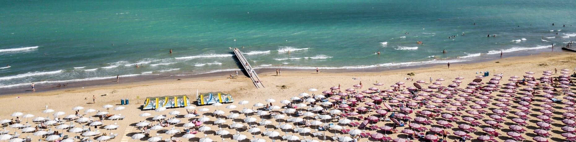 Spiaggia affollata con ombrelloni bianchi, sedie rosse e mare blu-verde, in una giornata calda e soleggiata.