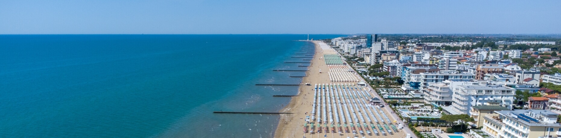 Vista panoramica di una città costiera con spiaggia, ombrelloni, lettini e edifici bianchi sotto un cielo blu.
