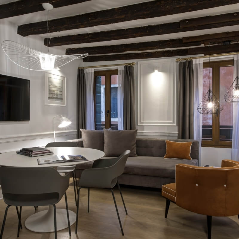 Modern living room with a white dining table, gray and orange chairs, gray couch, pendant lights, and warm wood beams.