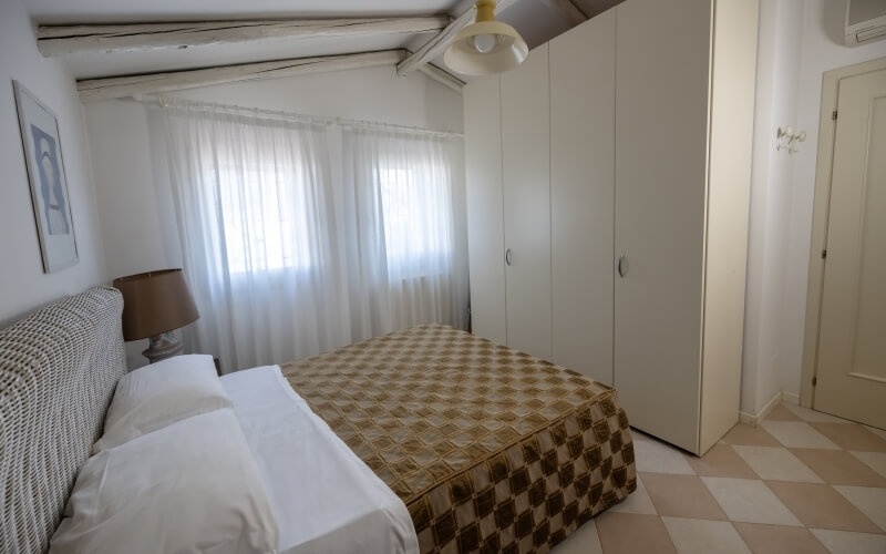 Camera da letto con letto a muro, armadio a cinque porte, lampada, finestra con tende bianche e soffitto in legno.